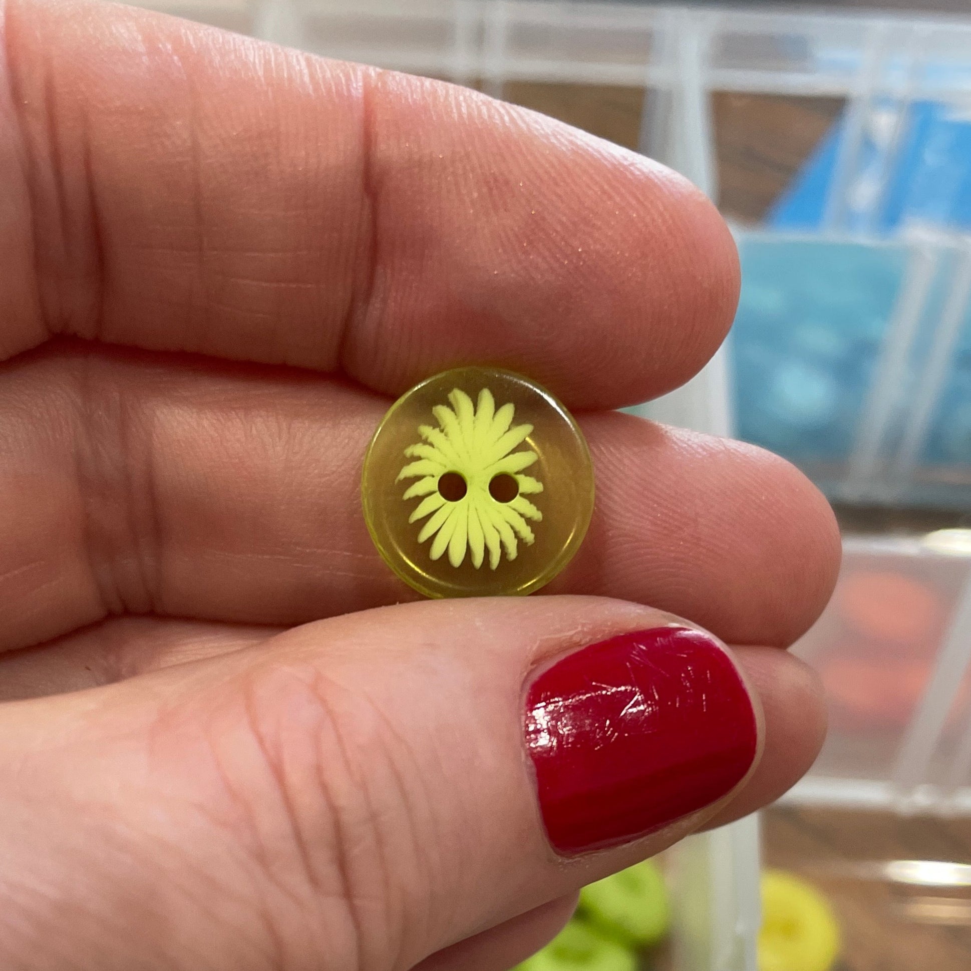 Hand holding a yellow button with a decorative center, against a background of button storage containers.