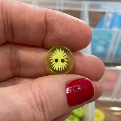 Hand holding a yellow button with a decorative center, against a background of button storage containers.