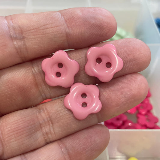 Pink flower-shaped buttons on a hand with a container of colorful buttons in the background.