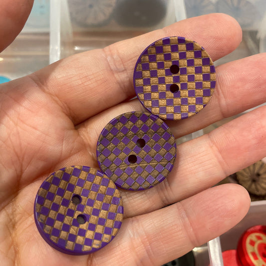 Three purple checkered buttons held in a hand with a container of buttons in the background.