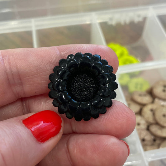 Black decorative item held between fingers with a container of buttons in the background