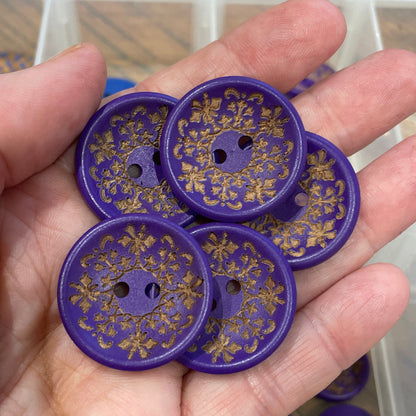 Hand holding purple buttons with floral patterns, with a clear container of more buttons in the background.