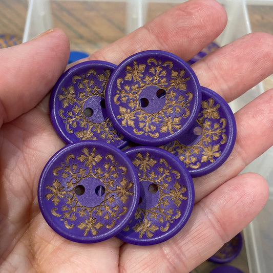 Hand holding purple buttons with floral patterns, with a clear container of more buttons in the background.