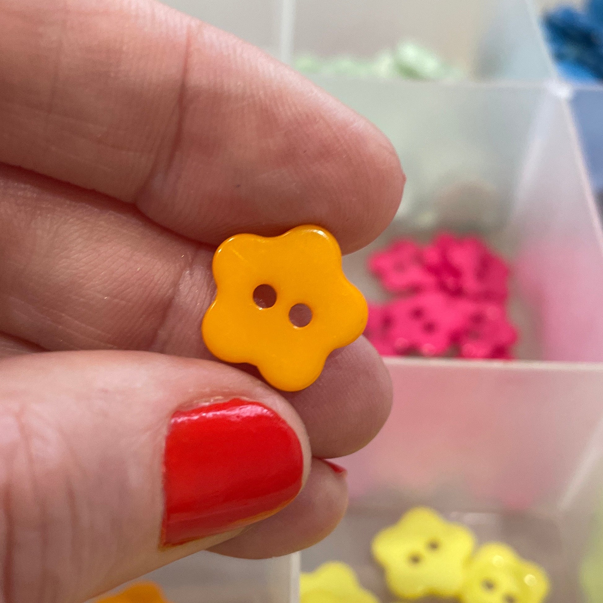 Hand holding a yellow flower-shaped button with a background of more buttons in a container.