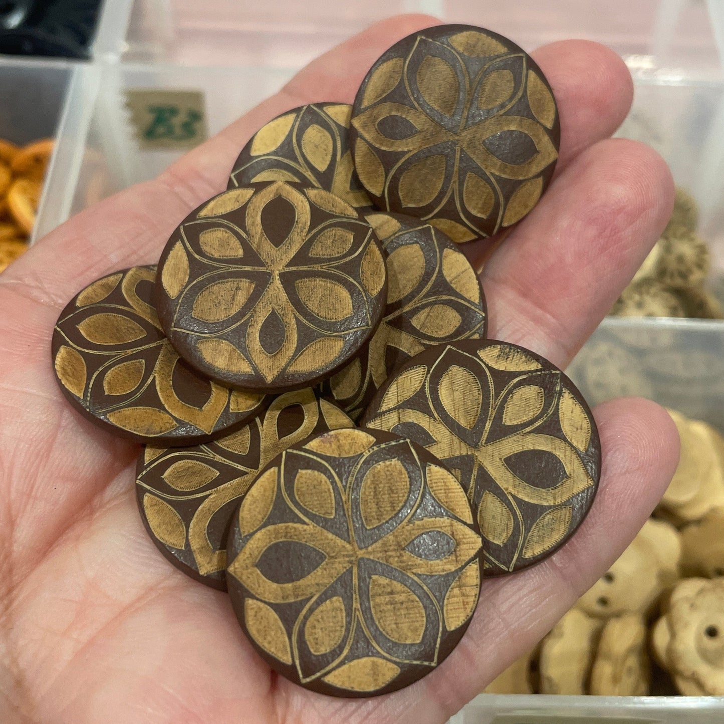Hand holding wooden buttons with intricate designs, blurred background of a storage box with more items.