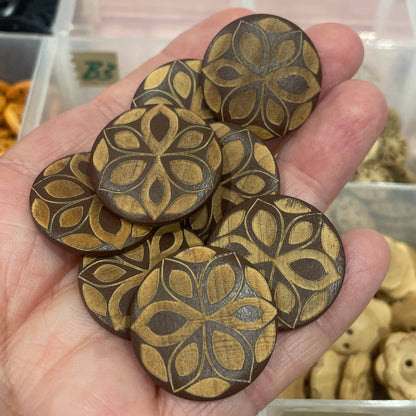 Hand holding wooden buttons with intricate designs, blurred background of a storage box with more items.
