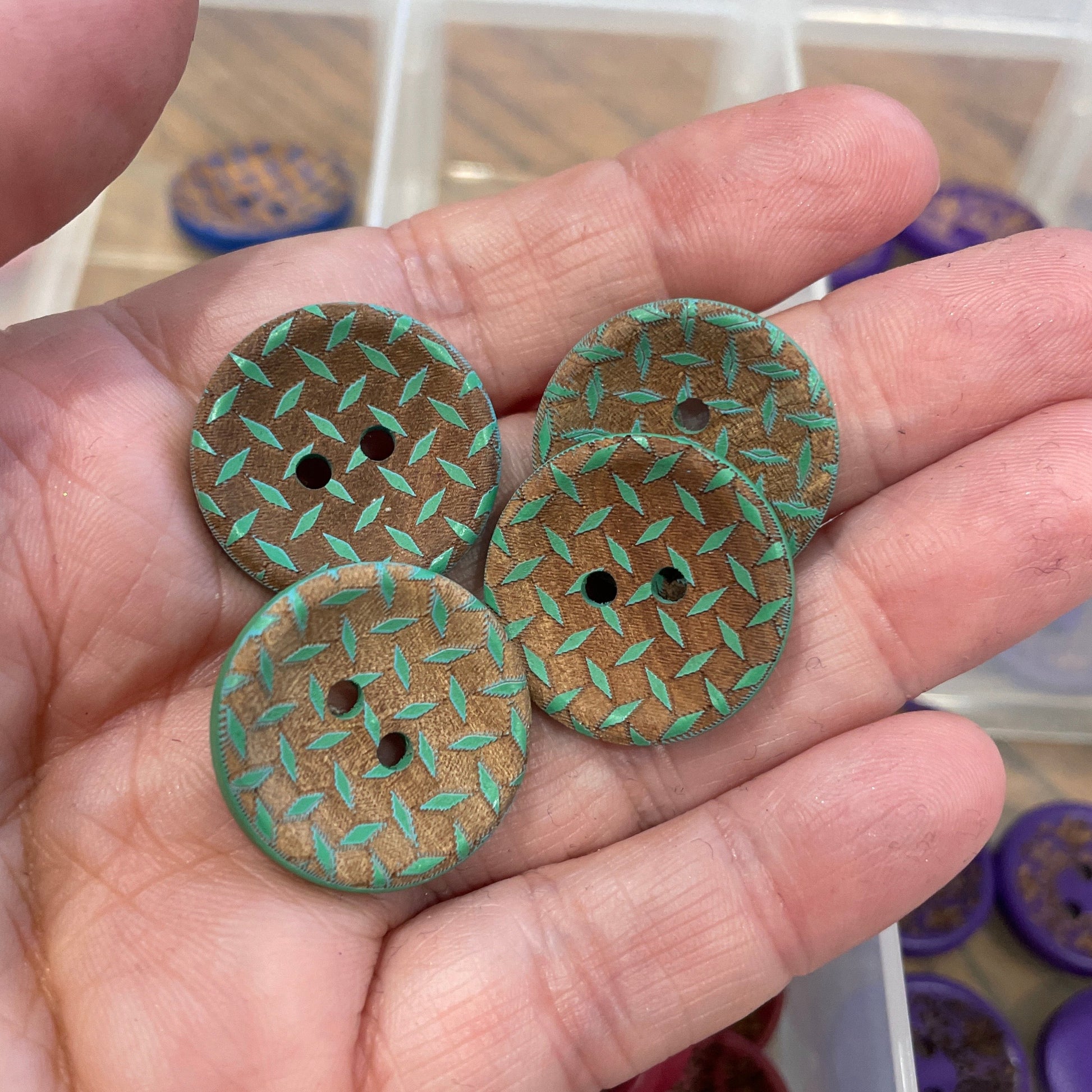 Three green textured buttons held in a hand with a container of buttons in the background.