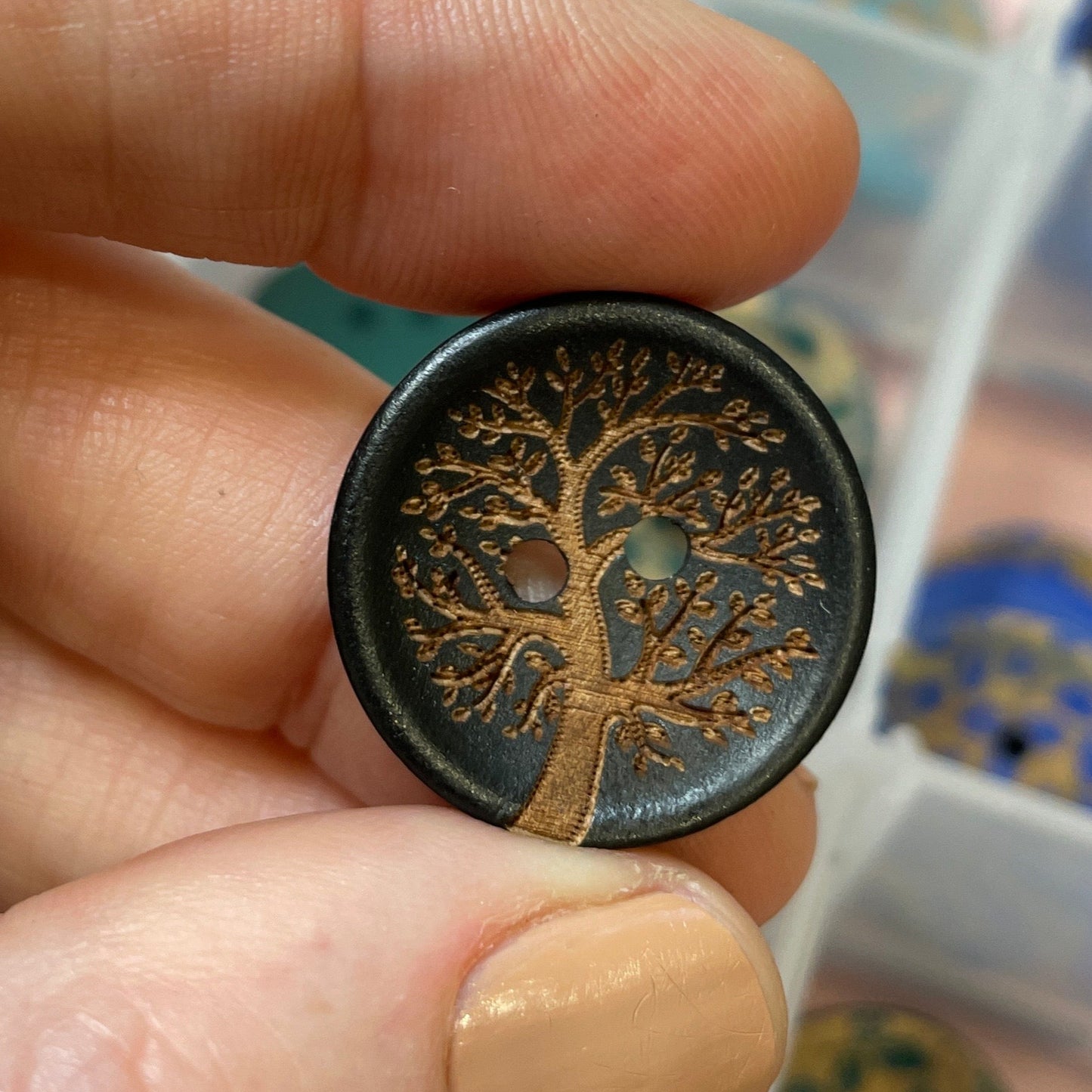 Hand holding a decorative button with intricate design, against a blurred background of a jewelry box.