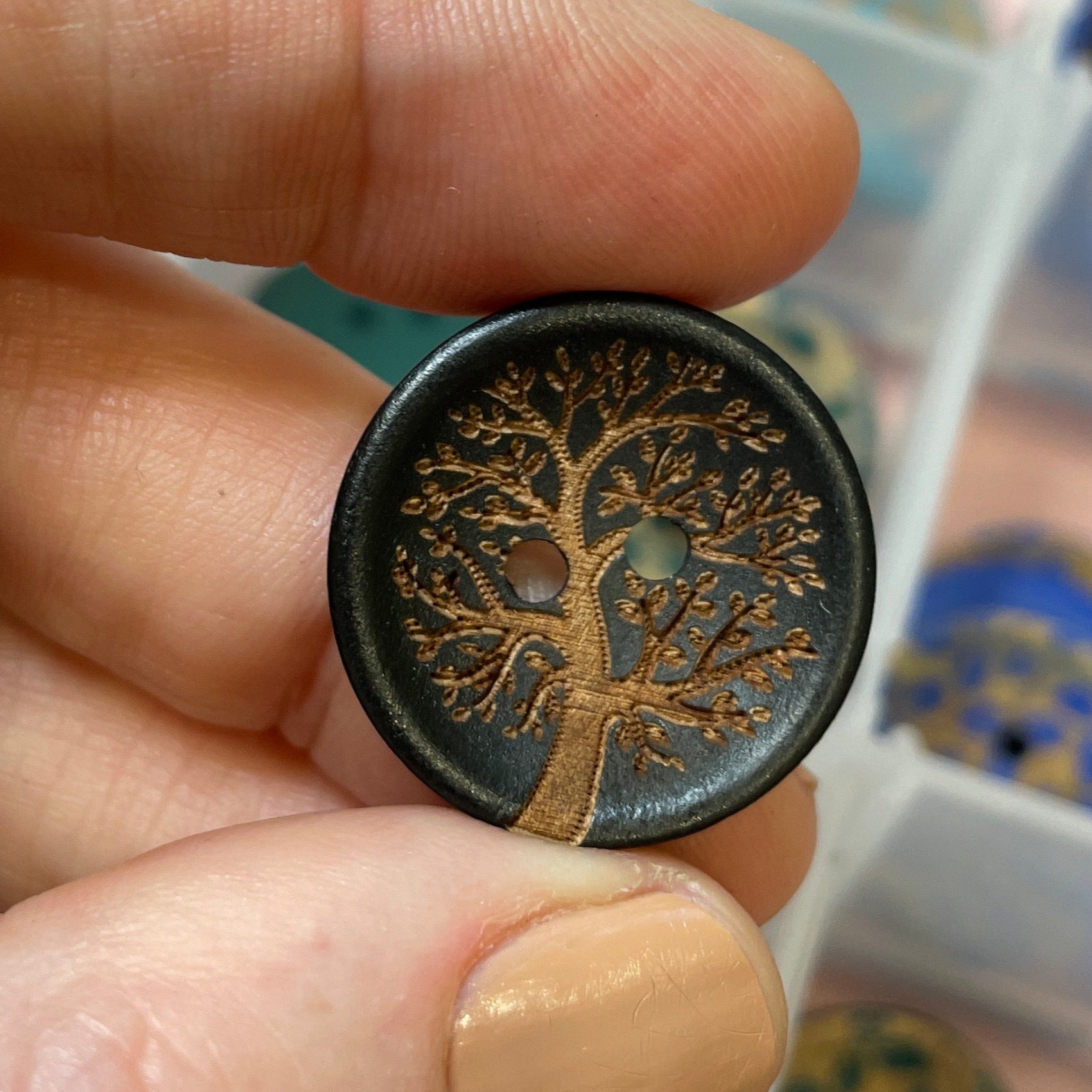 Hand holding a decorative button with intricate design, against a blurred background of a jewelry box.