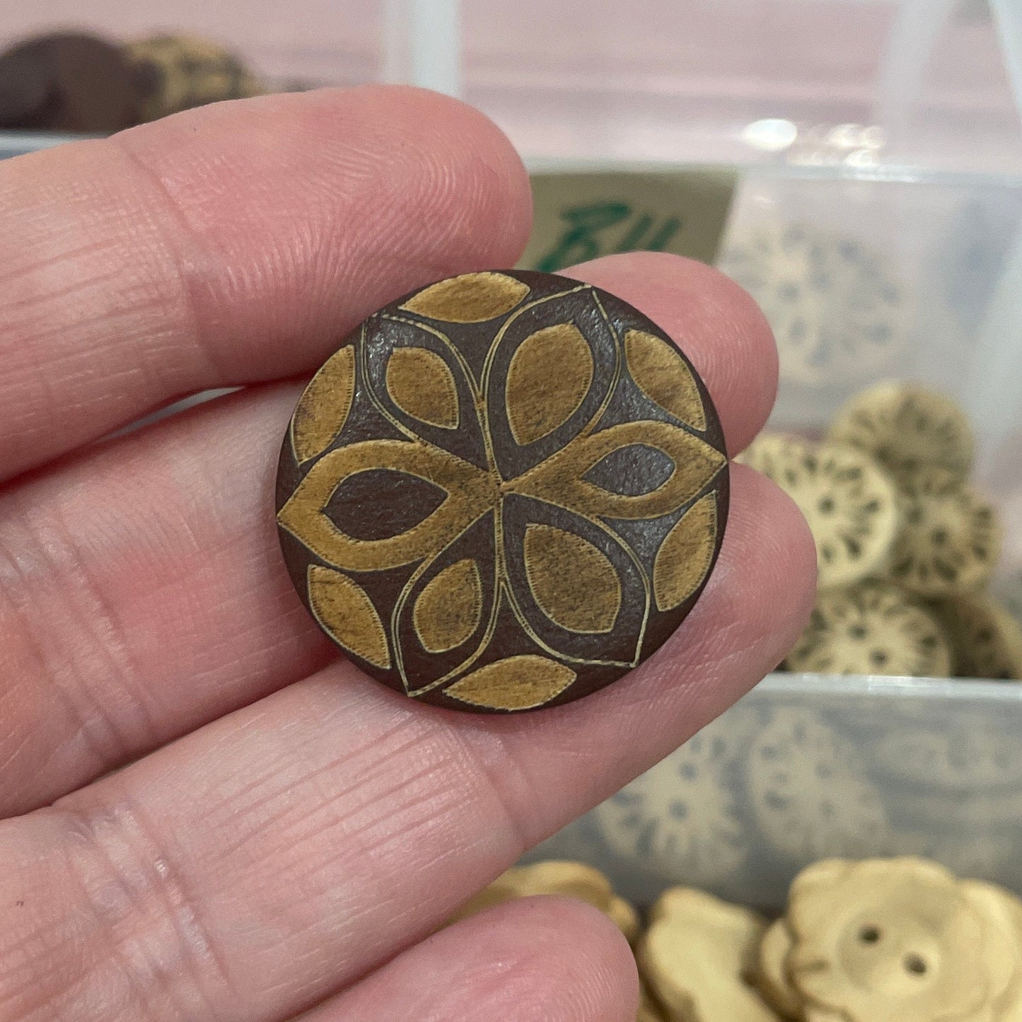 Hand holding a decorative button with a detailed design, against a background of similar buttons in a container.