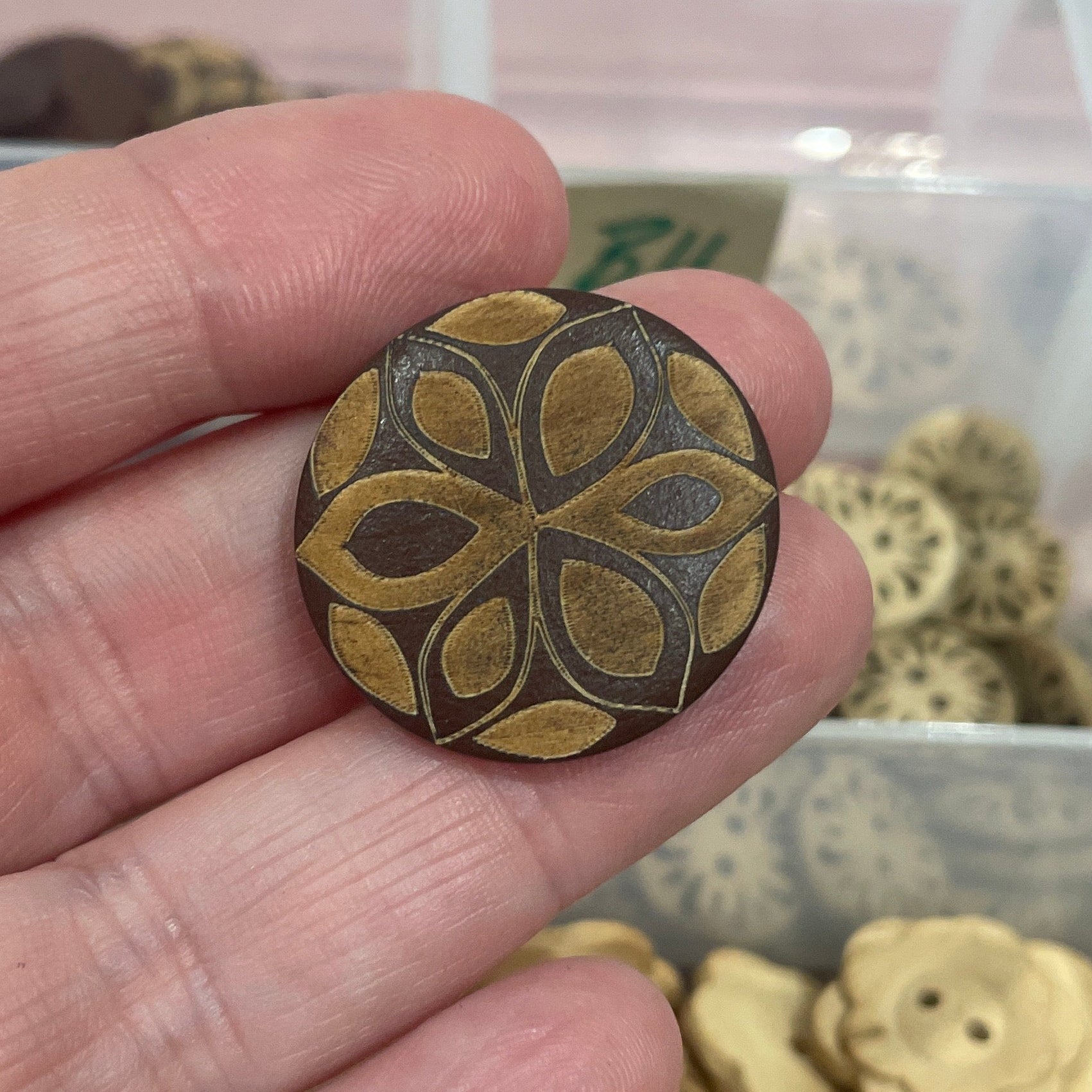 Hand holding a decorative button with a detailed design, against a background of similar buttons in a container.