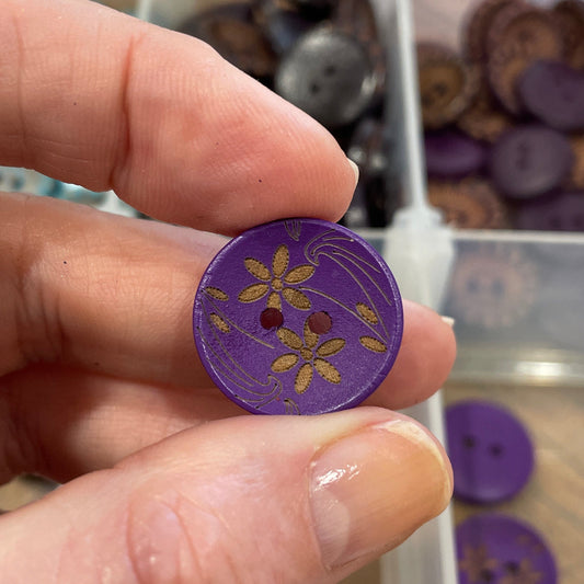 Hand holding a purple button with floral design in front of a container of buttons.