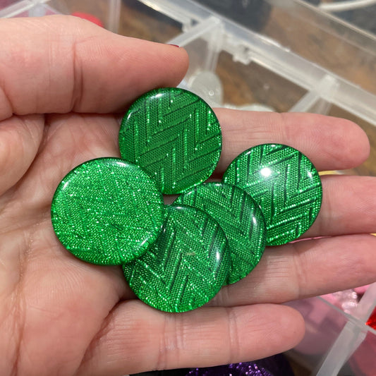 Hand holding green circular objects with a container of various colored objects in the background