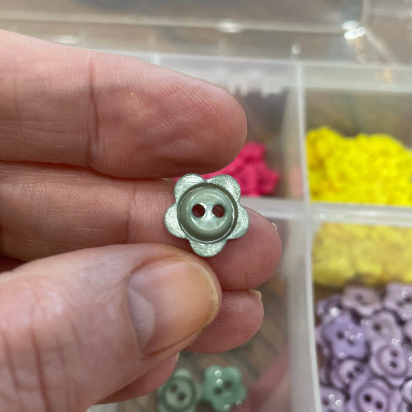 Hand holding a silver button with a clear container of assorted buttons in the background