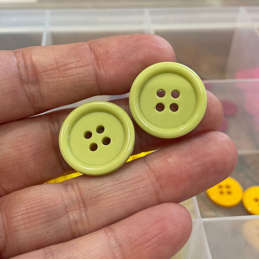 Two green buttons held between fingers with a blurred background of a storage box.