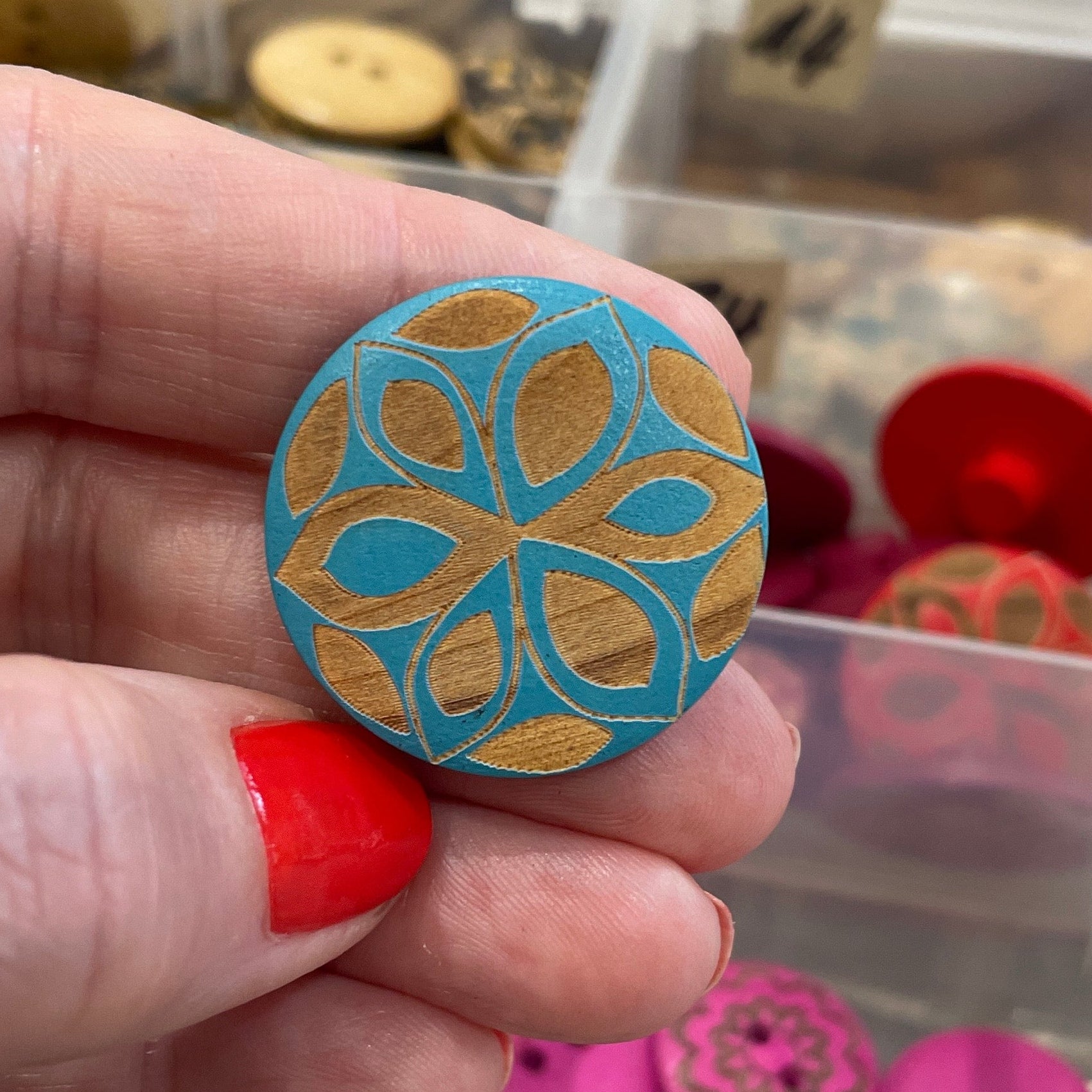 Hand holding a blue patterned button with a container of assorted buttons in the background.
