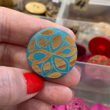 Hand holding a blue patterned button with a container of assorted buttons in the background.