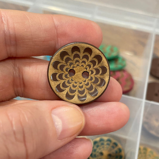 Hand holding a decorative wooden button with a detailed design, against a background of similar buttons in a container.