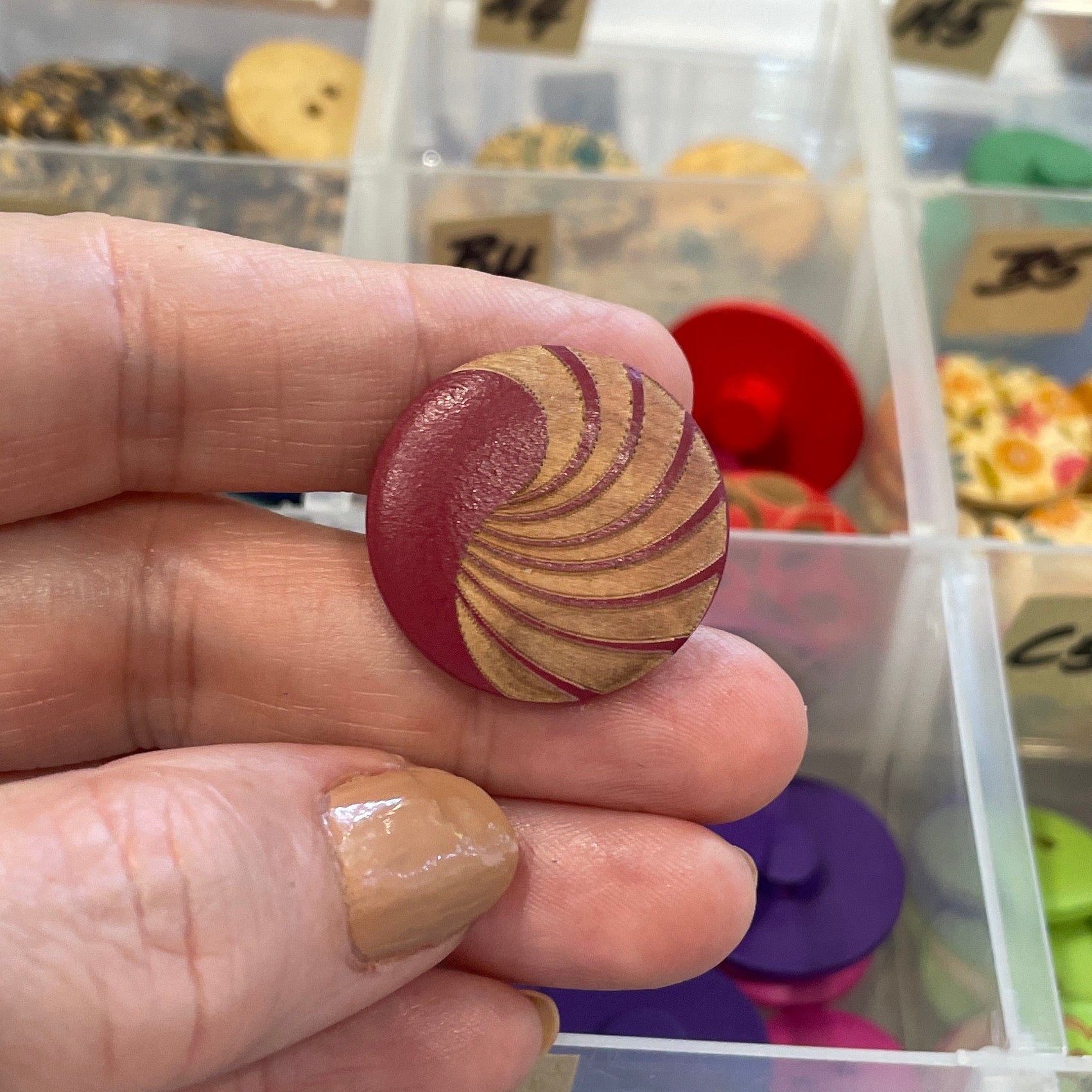 Hand holding a decorative button in front of a container filled with various buttons.