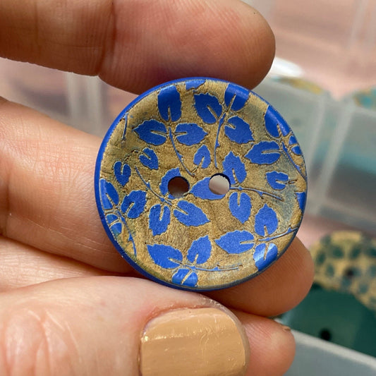 Hand holding a decorative button with blue floral pattern, surrounded by a container of buttons.
