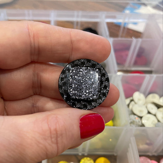 Hand holding a black glittery gemstone with a container of colorful gemstones in the background.