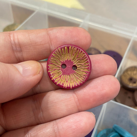 Hand holding a pink button with a blurred background of button assortment