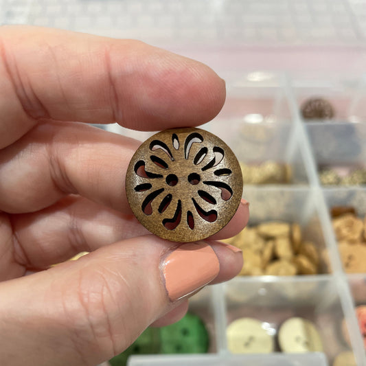 Hand holding a decorative brass button with intricate design, against a blurred background of a container with more buttons.