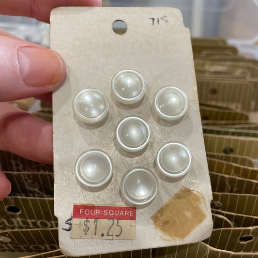 Card with pearl buttons held by a hand, with a container of similar buttons in the background.