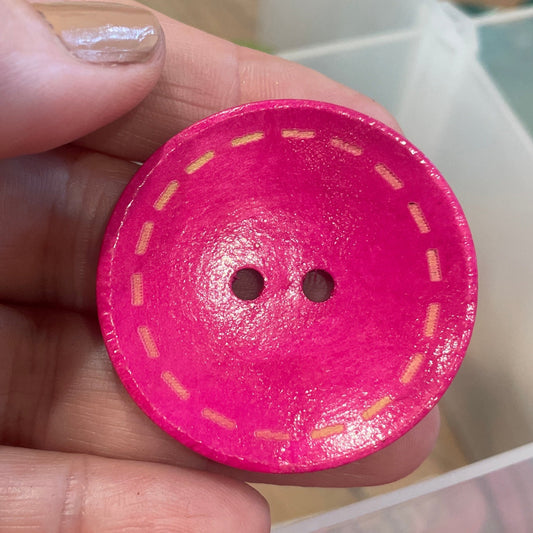 Pink button with yellow stitching held by a hand, with more buttons in the background.