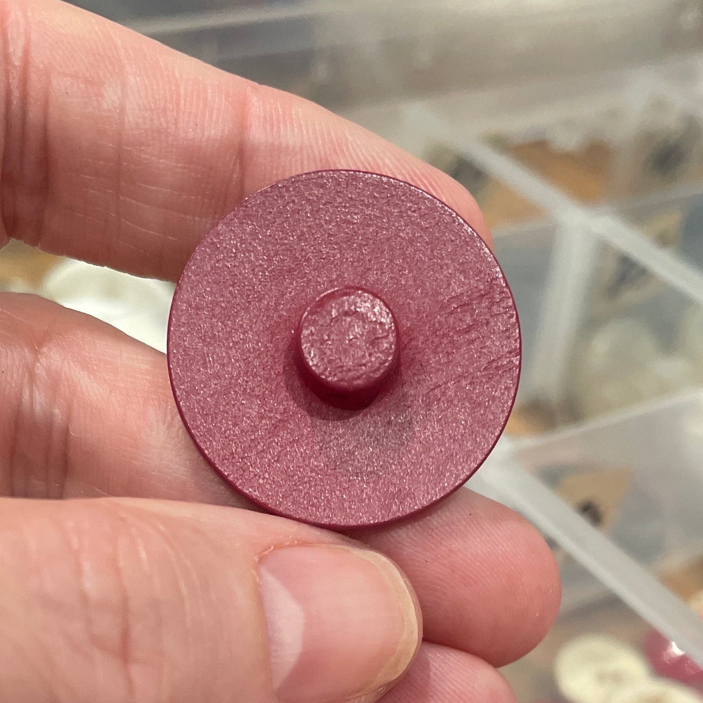 Hand holding a round button with a container of assorted colored objects in the background