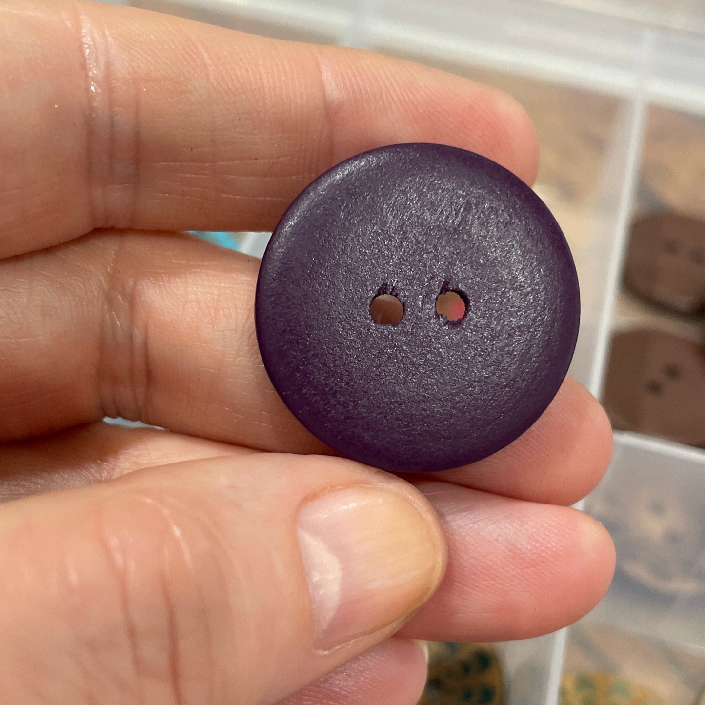 Hand holding a purple button with a container of various buttons in the background