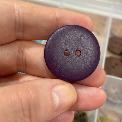 Hand holding a purple button with a container of various buttons in the background