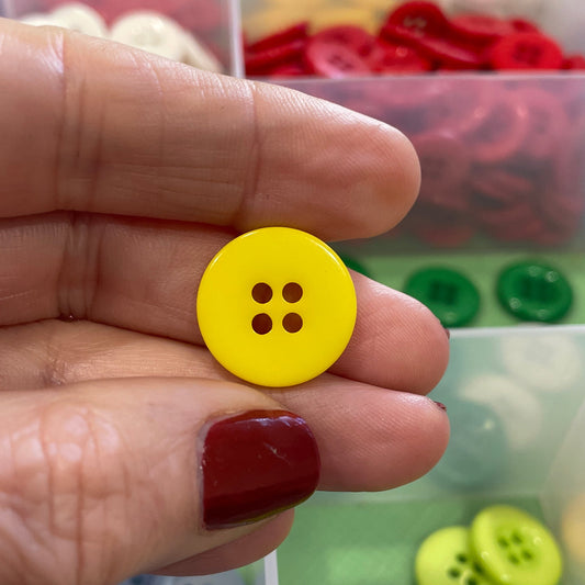 Hand holding a yellow button with four holes in front of a container of colorful buttons.