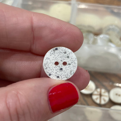 Hand holding a round, glittery button with a clear container of buttons in the background