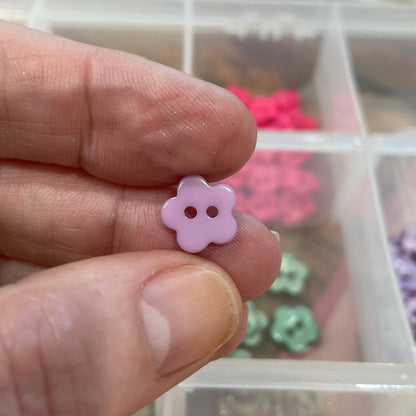 Hand holding a small pink flower-shaped object with a container of colorful buttons in the background.