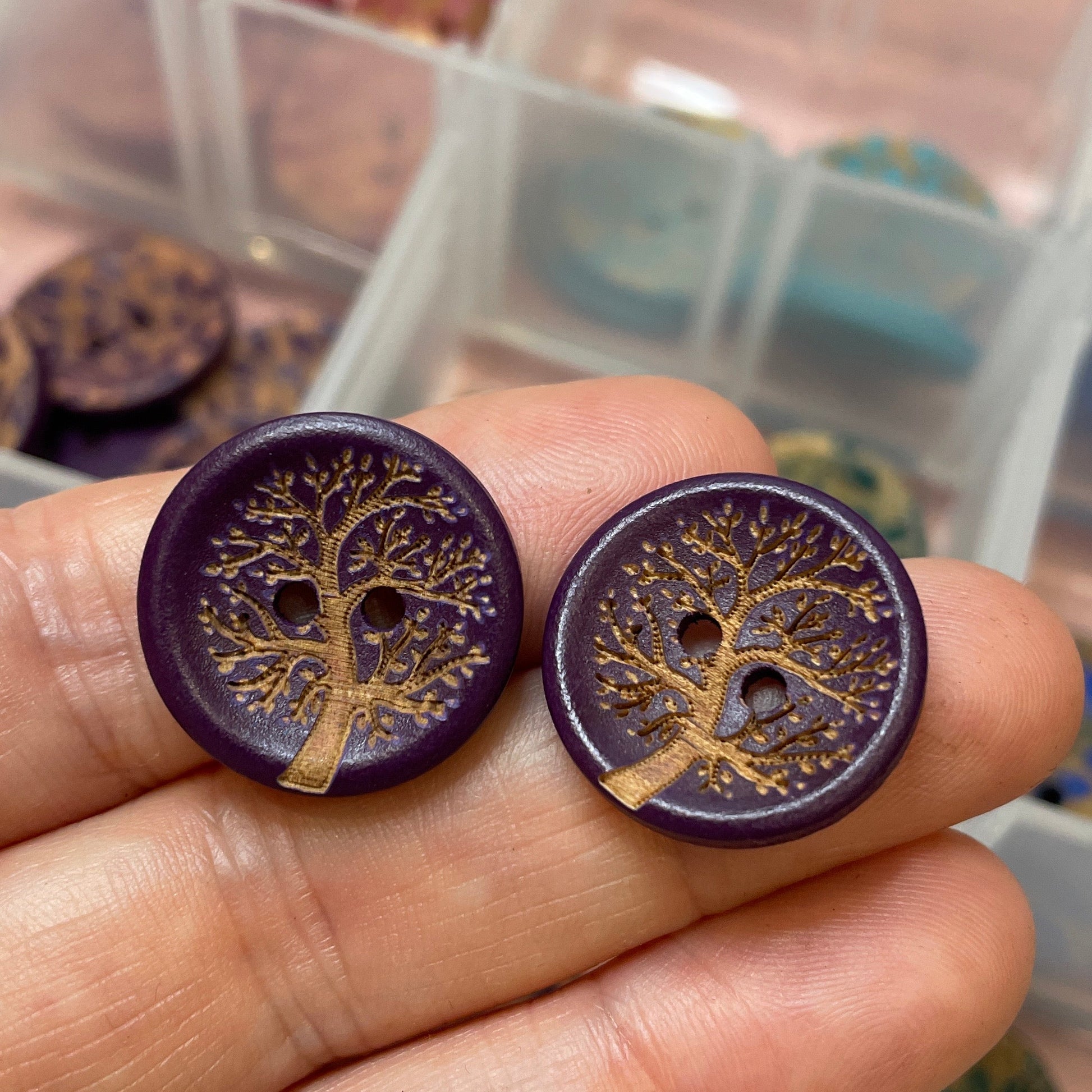 Purple buttons with tree design held by a hand, with a container of similar buttons in the background.
