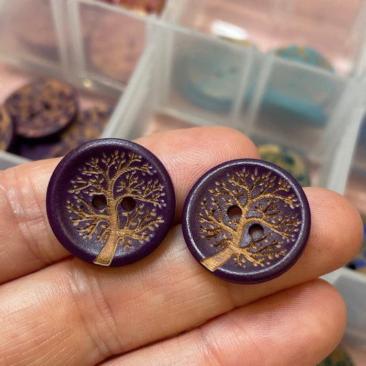 Purple buttons with tree design held by a hand, with a container of similar buttons in the background.