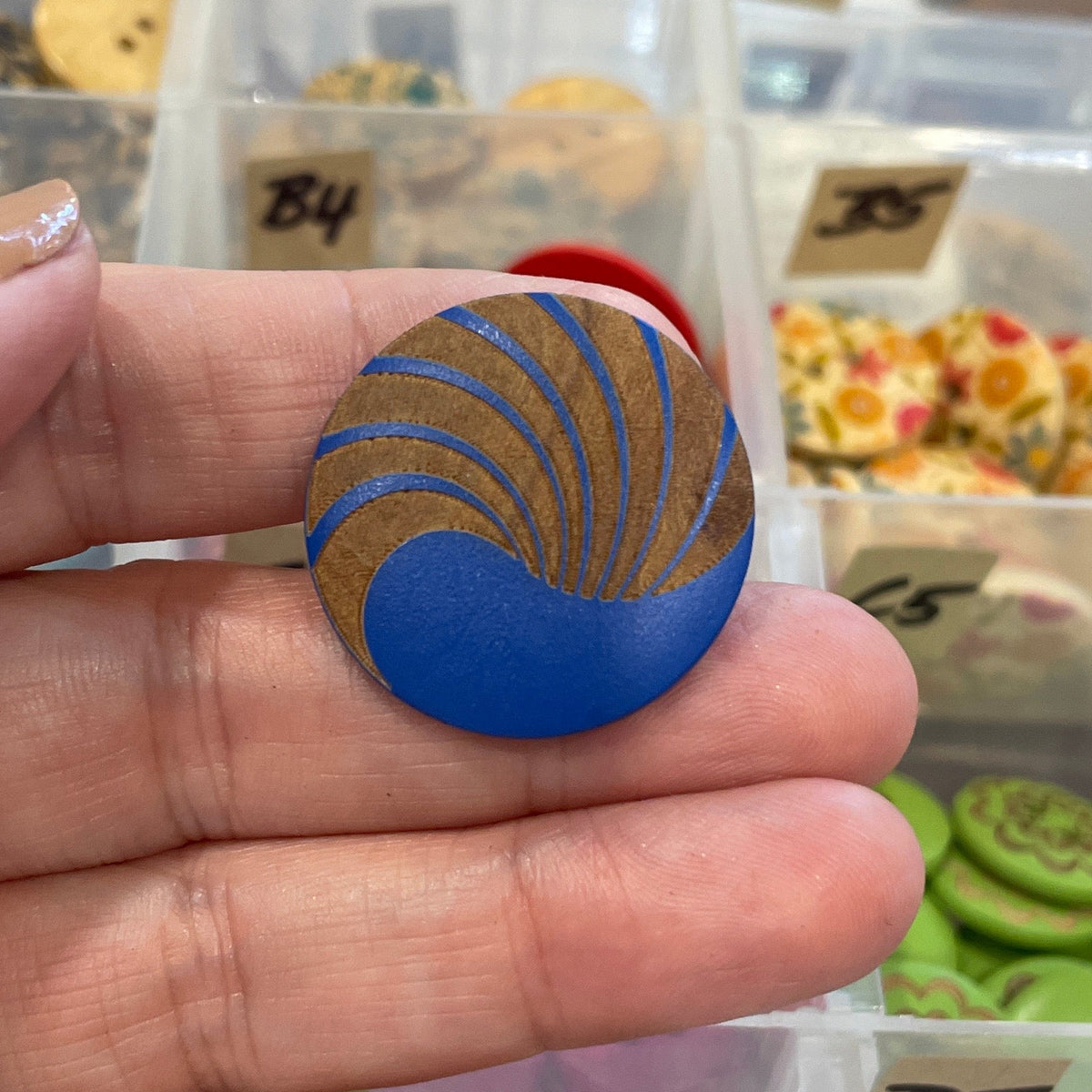 Hand holding a blue and gold button with a background of various buttons in a clear container.