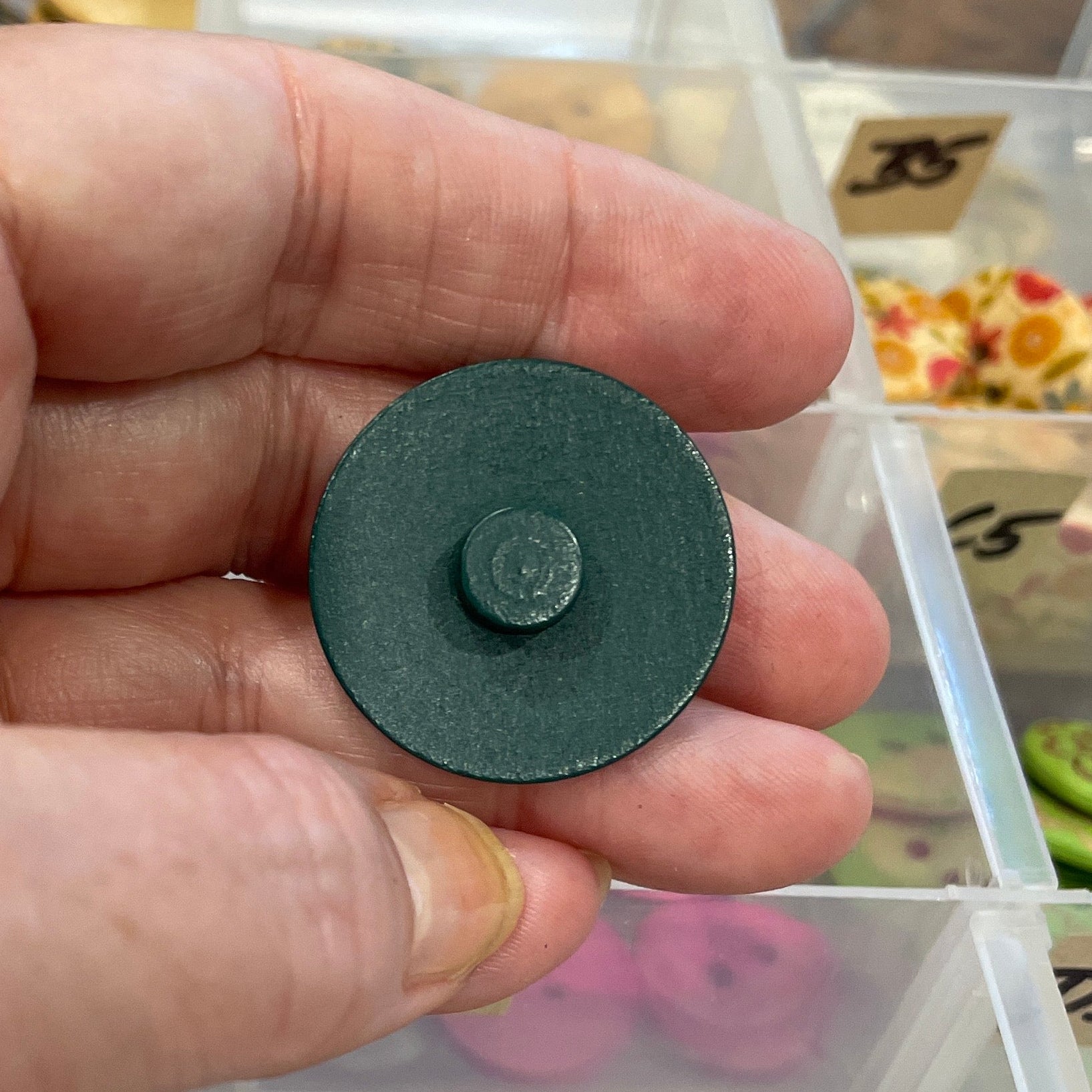 Hand holding a black button in front of a clear container with various buttons.
