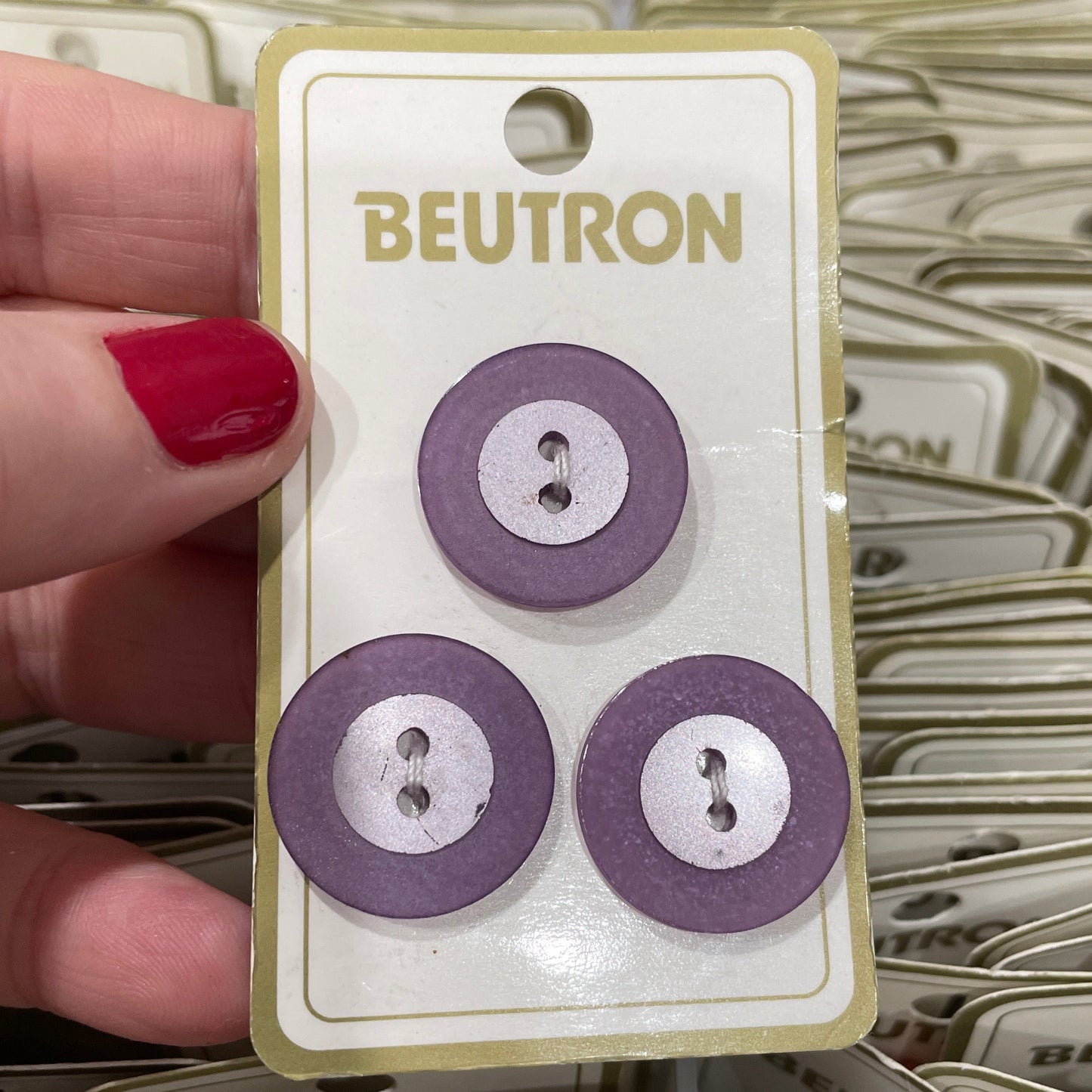 Three purple buttons on a Beutron card held by a hand with red nail polish.