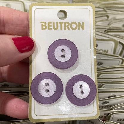 Three purple buttons on a Beutron card held by a hand with red nail polish.