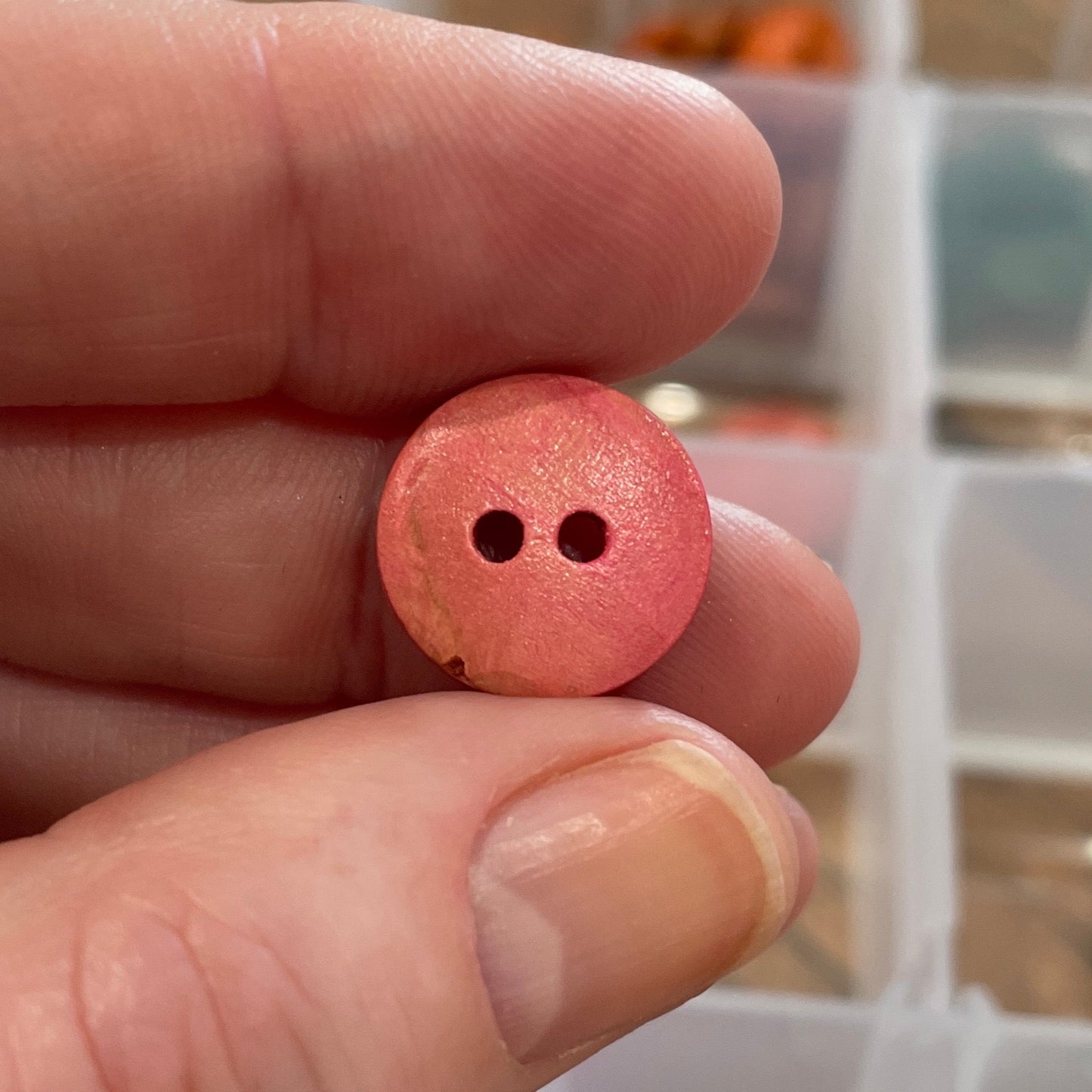 Hand holding a pink button with two holes, with a container of buttons in the background.