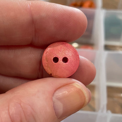 Hand holding a pink button with two holes, with a container of buttons in the background.