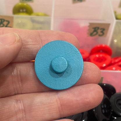 Hand holding a blue button with a container of assorted buttons in the background