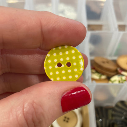 Hand holding a yellow button with white polka dots in front of a container of buttons.