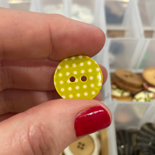 Hand holding a yellow button with white polka dots in front of a container of buttons.
