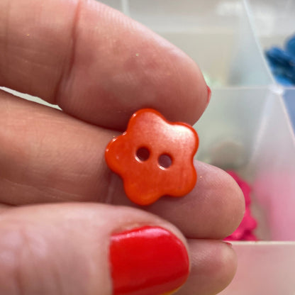Hand holding an orange flower-shaped button with a container of colorful buttons in the background.