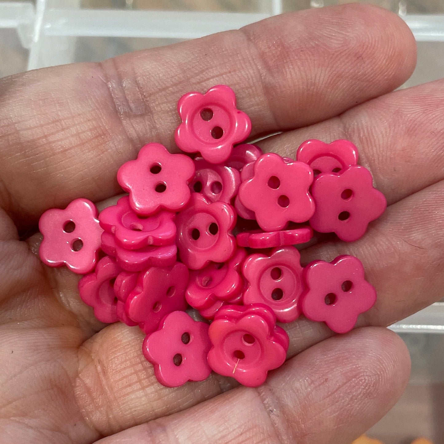 Hand holding pink flower-shaped objects with a clear container of assorted small items in the background.
