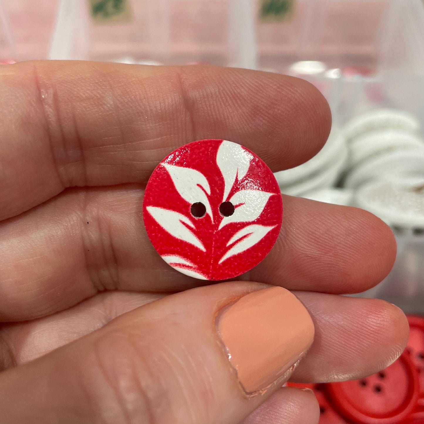 Red and white button held between fingers with blurred container of buttons in the background