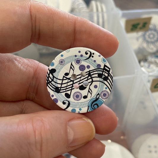 Hand holding a decorative button with musical notes in front of a container of buttons.